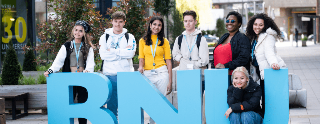 Six diverse students pose behind large blue letters on a campus plaza, smiling at the camera.