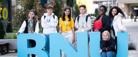 Six diverse students pose behind large blue letters on a campus plaza, smiling at the camera.