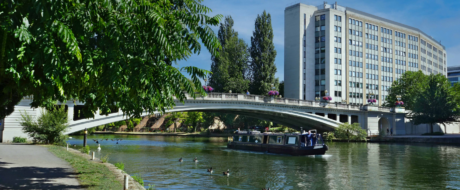 A river scene with a white arched bridge, a canal boat passing beneath, and a tall modern building in the background as trees line the bank.