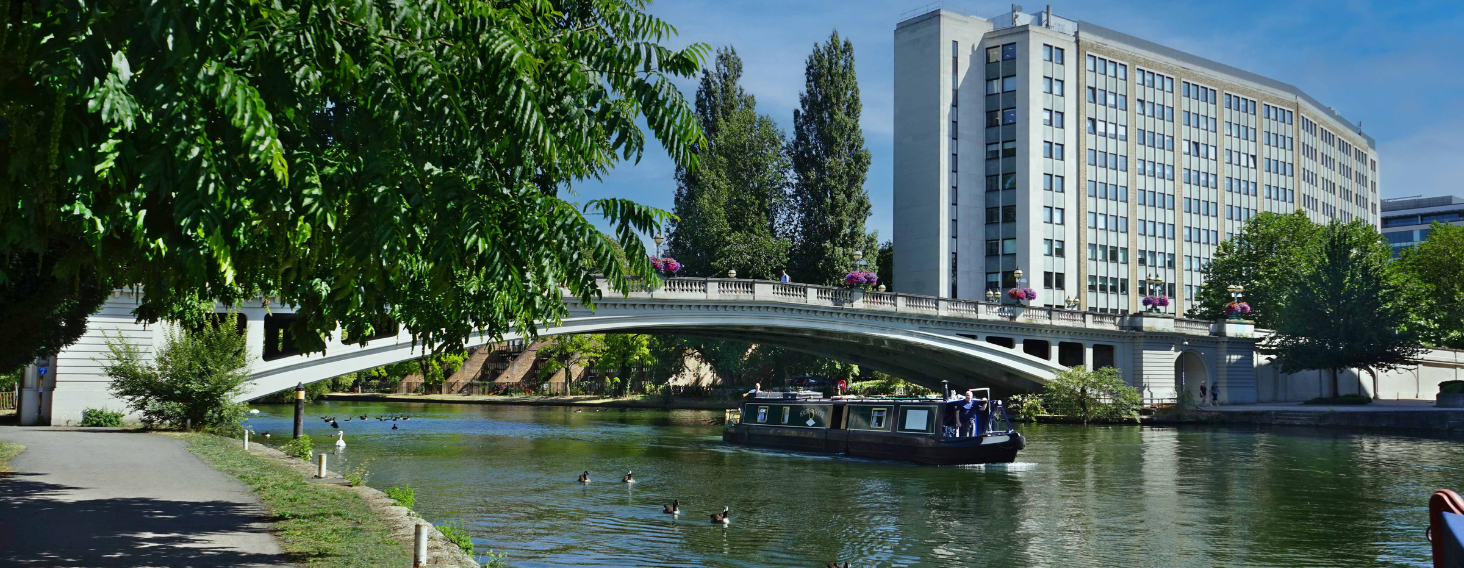 A river scene with a white arched bridge, a canal boat passing beneath, and a tall modern building in the background as trees line the bank.