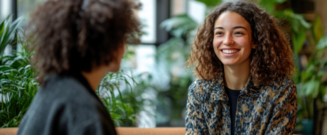 Two people chatting in a bright, plant-filled space; woman with curly hair smiling at the other person.