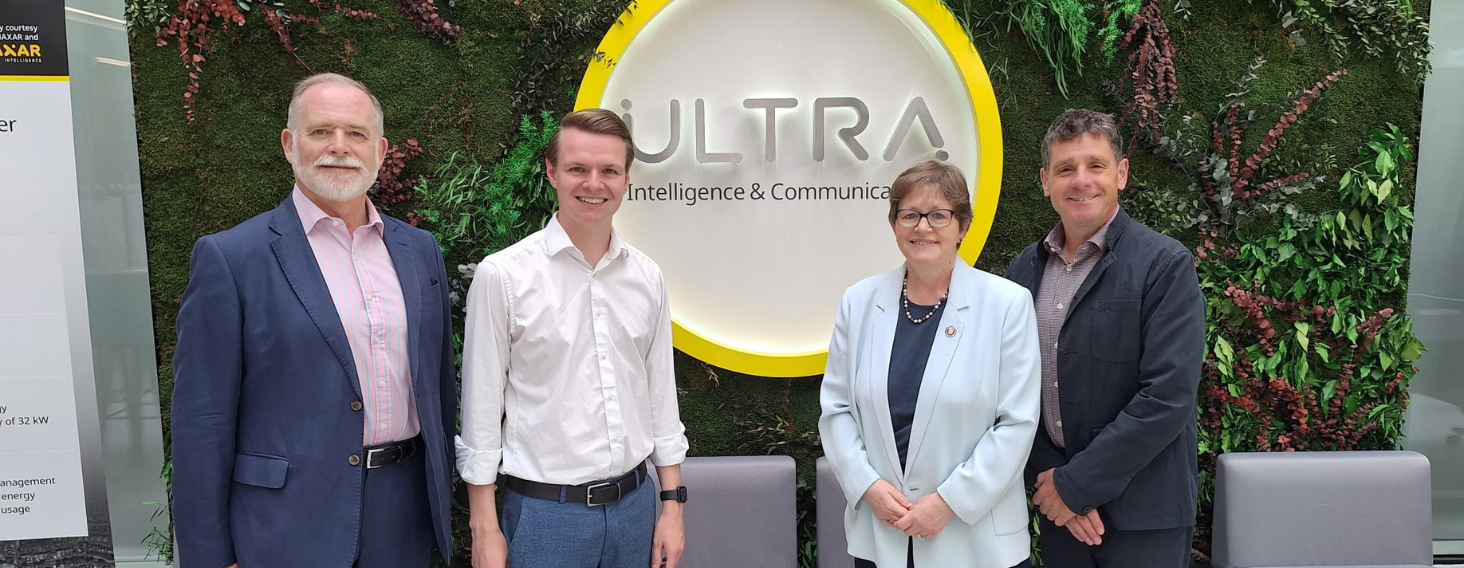 Four professionals stand side by side in front of a circular ULTRA sign on a green living wall, posing for a group photo in a corporate setting.