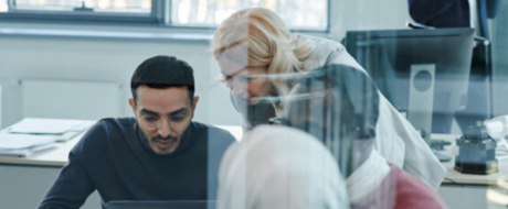 Three colleagues collaborate over a laptop at a bright office desk, with a glass partition in the foreground.