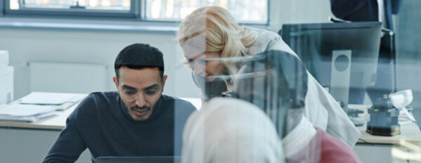 Three colleagues collaborate over a laptop at a bright office desk, with a glass partition in the foreground.