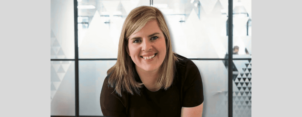 Smiling blonde woman in a dark shirt leaning forward in a bright office with frosted glass panels in the background