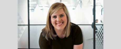 Smiling blonde woman in a dark shirt leaning forward in a bright office with frosted glass panels in the background