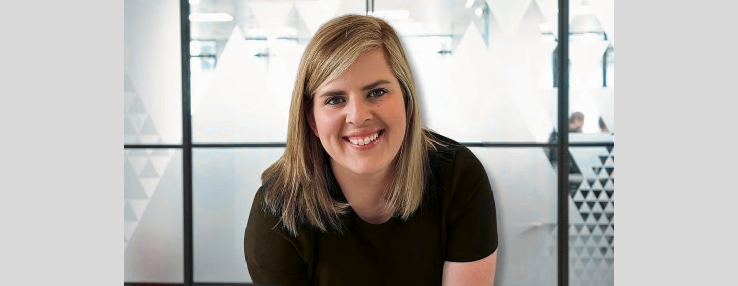 Smiling blonde woman in a dark shirt leaning forward in a bright office with frosted glass panels in the background