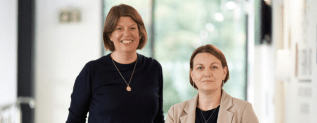 Two smiling women in a bright office corridor, one in a black top with a gold pendant, the other in a beige blazer taking a friendly photo together.