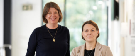 Two smiling women in a bright office corridor, one in a black top with a gold pendant, the other in a beige blazer taking a friendly photo together.
