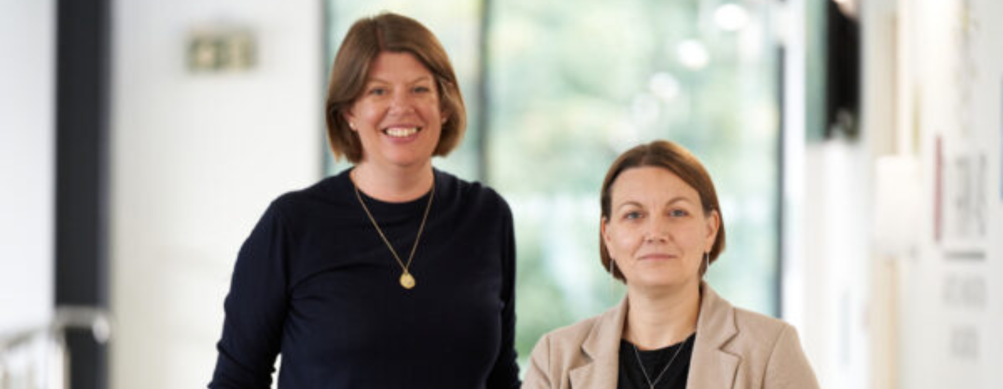 Two smiling women in a bright office corridor, one in a black top with a gold pendant, the other in a beige blazer taking a friendly photo together.