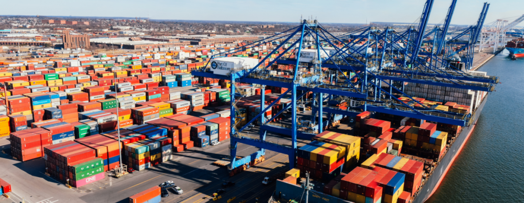 Large container port with colorful stacked shipping containers and tall blue gantry cranes over a docked cargo ship area