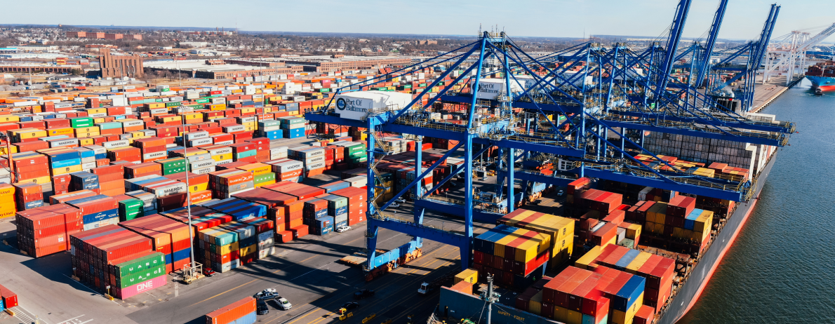 Large container port with colorful stacked shipping containers and tall blue gantry cranes over a docked cargo ship area