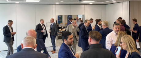 Group of professionals in business attire mingle and chat at a modern office reception with coffee cups and networking in the background.