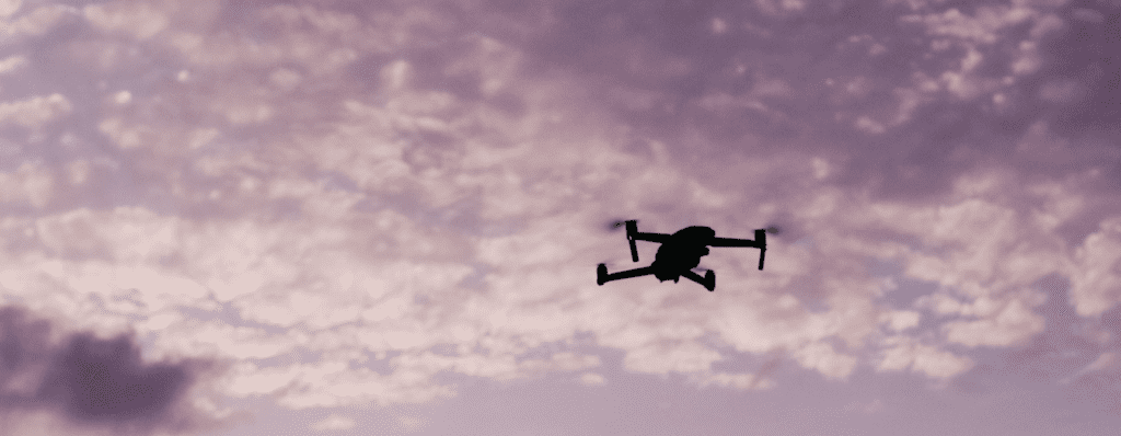Silhouette of a quadcopter drone flying against a pink-purple sunset with scattered clouds.