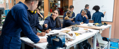 Group of students in blue coveralls collaborate at a workshop table with tools and a whiteboard in the background, smiling and taking notes amid hands-on learning.