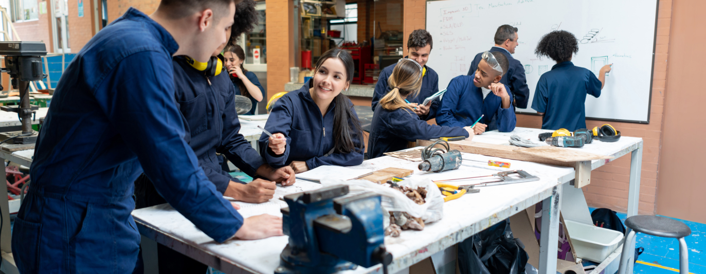 Group of students in blue coveralls collaborate at a workshop table with tools and a whiteboard in the background, smiling and taking notes amid hands-on learning.