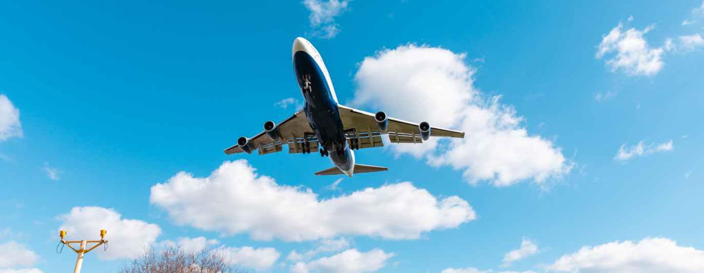 Low-angle view of a large commercial airliner flying overhead against a blue sky with clouds; airport light pole visible bottom left.