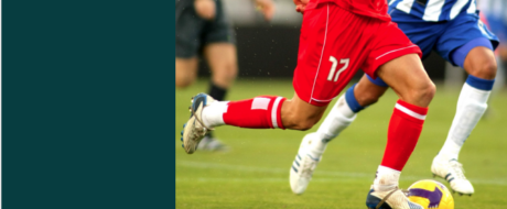 Action shot of soccer players' legs contending for the ball on a green field; a player in red kit (number 17) is near the ball.
