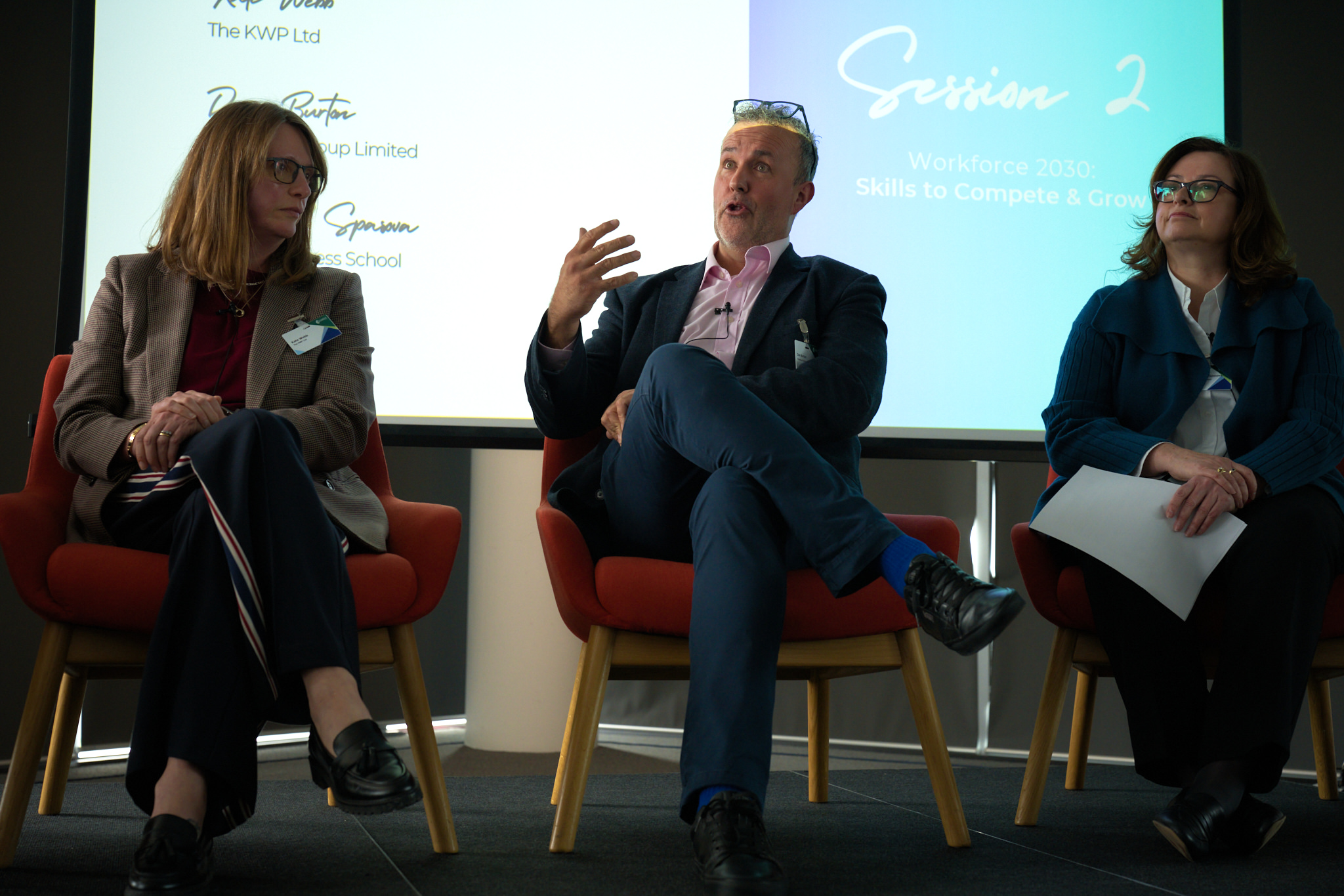 Three panelists seated on red chairs during a discussion, with a bright presentation screen behind them.