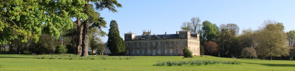 Historic stone manor in a sunny park, surrounded by a well-kept lawn and tall trees under a clear blue sky.