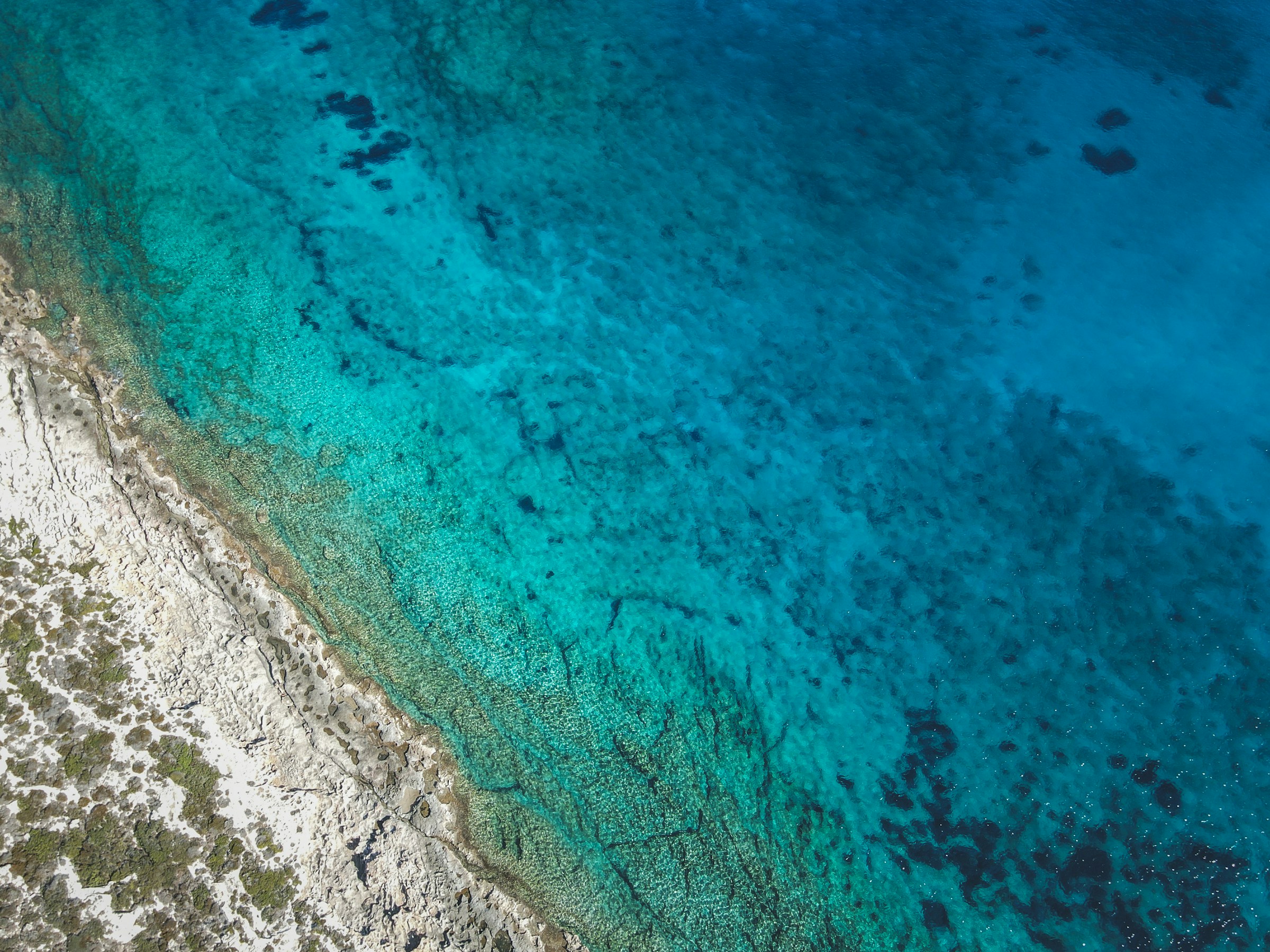 Aerial view of turquoise water along a rocky coastline, with clear shallow areas near shore.
