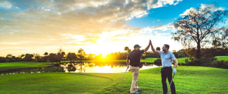 Two golfers on a sunlit golf course celebrate with a high-five after a shot at sunset.