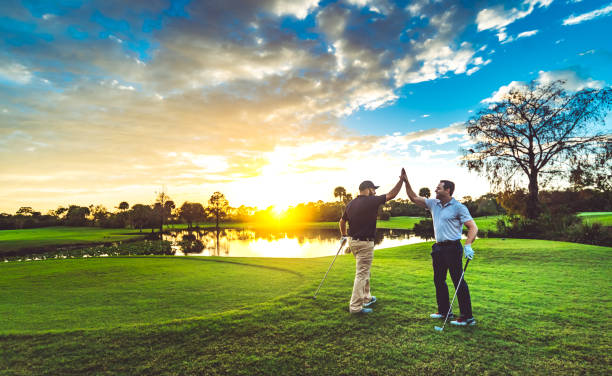 Two golfers on a sunlit golf course celebrate with a high-five after a shot at sunset.