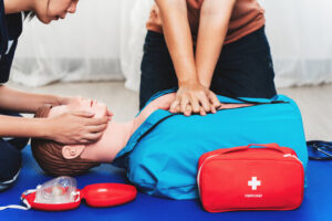 Two people practice CPR on a lifelike manikin on a blue mat; one person tilts the head while the other presses on the chest, with a red first aid kit nearby.