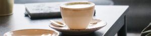 White ceramic coffee cup with frothy top on a matching saucer and a spoon, set on a dark wooden table (background is blurred).