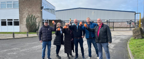 Eight people stand outside an industrial building, toasting with orange cups to celebrate together.