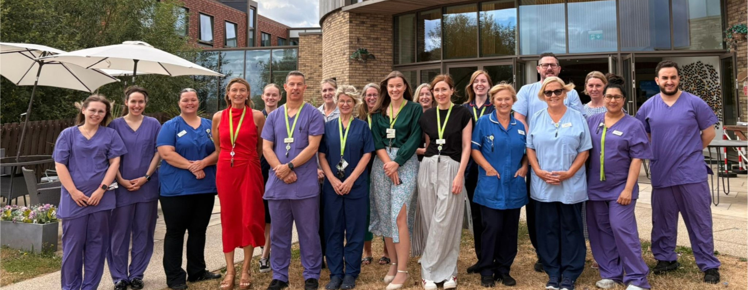 NEW - News item size 63 | Thames Valley Chamber of Commerce Group of healthcare workers in purple, blue, and red scrubs posing outdoors in front of a brick building with umbrellas nearby.