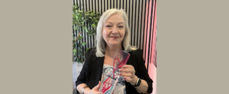 Smiling woman in a black blazer holding a glass award labeled WINNER, standing beside a striped wall and plants.