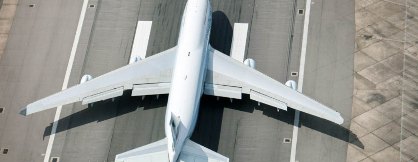 NEW - News item size 66 | Thames Valley Chamber of Commerce Top-down view of a large white commercial airplane aligned on a gray runway, wings spread wide and shadowed on the tarmac