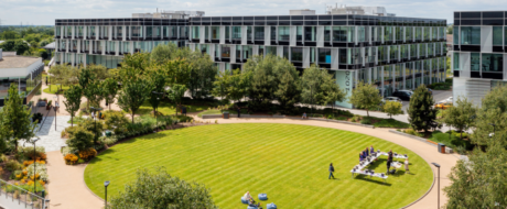 Aerial view of a modern campus with a circular green lawn, tree-filled courtyard, and glass-office buildings; people walk and sit at picnic tables.