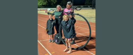 Girl in a navy raincoat stands on a running track with a large-wheeled bike-like device; two adults and another child in jackets smile and pose nearby on a sunny day, suggesting a family cycling event.