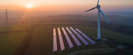Sunset over a renewable energy site with solar panels and a wind turbine on a hillside.