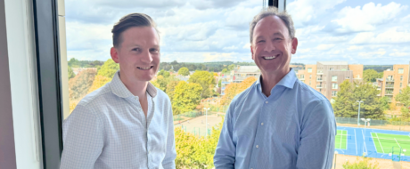 Two men in light shirts stand by a window, smiling, with trees and blue tennis courts visible outside behind them.