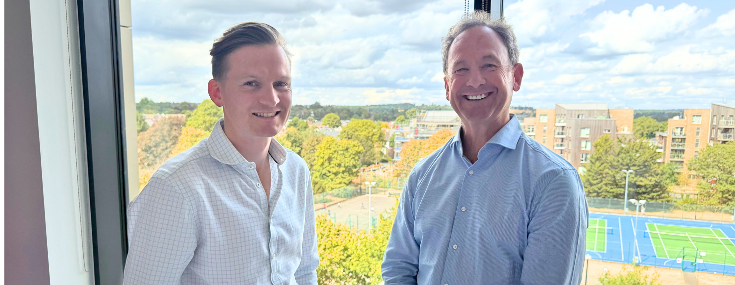 Two men in light shirts stand by a window, smiling, with trees and blue tennis courts visible outside behind them.