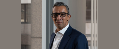 Professional man with short gray hair and glasses in a navy suit, standing by a window in a modern office environment