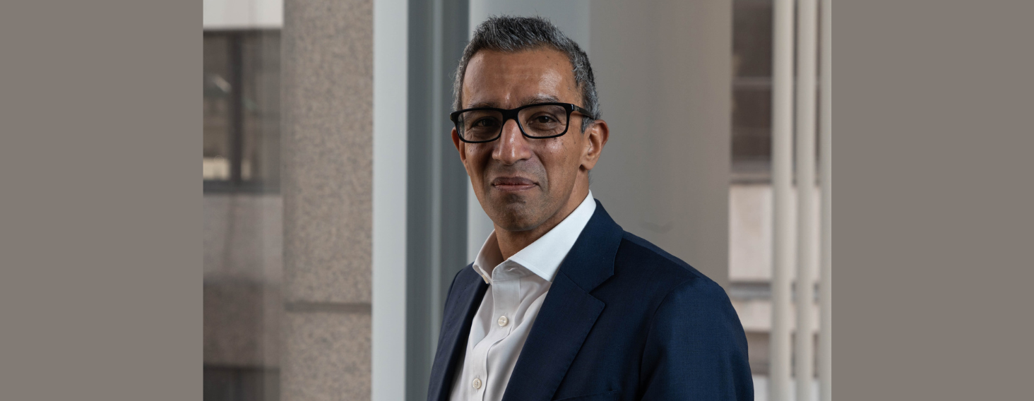 Professional man with short gray hair and glasses in a navy suit, standing by a window in a modern office environment