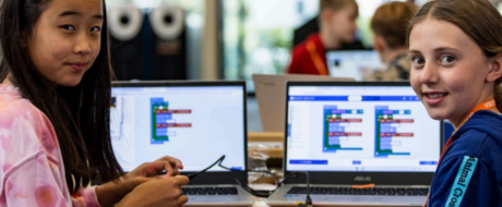 Two girls work on laptops at a coding workshop, with block-based programming visible on their screens