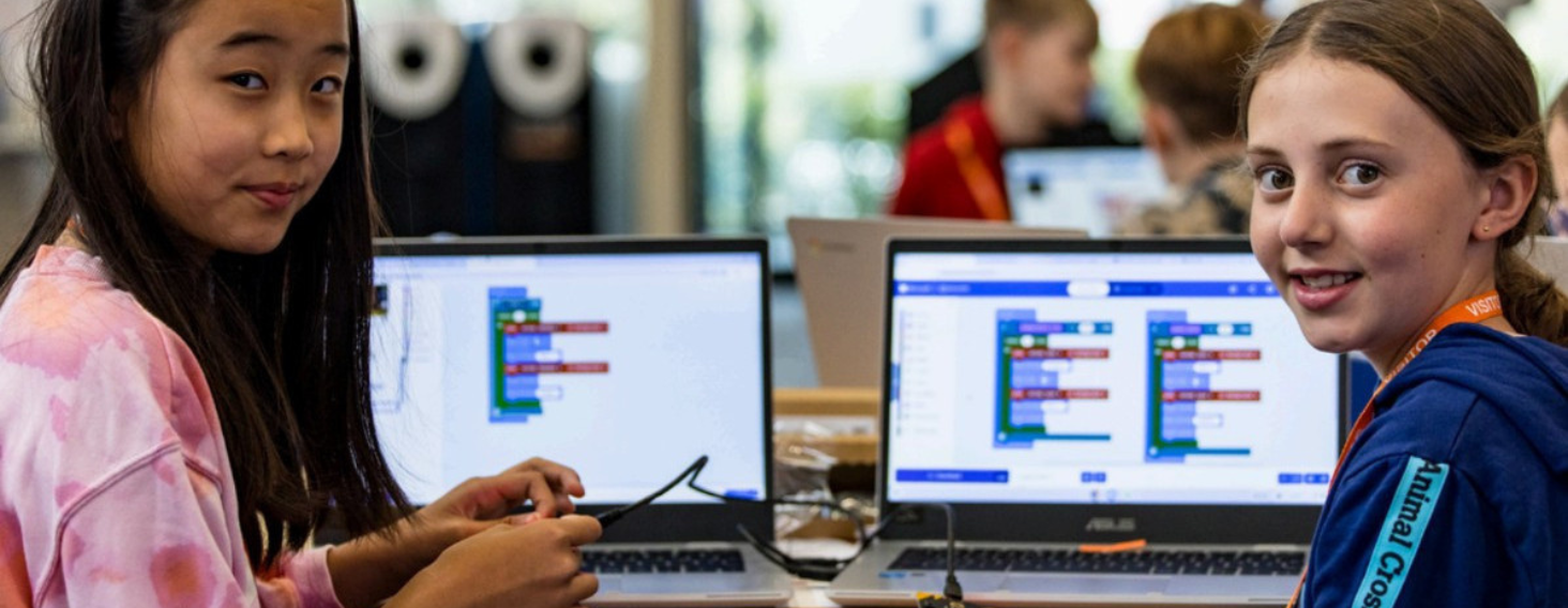Two girls work on laptops at a coding workshop, with block-based programming visible on their screens