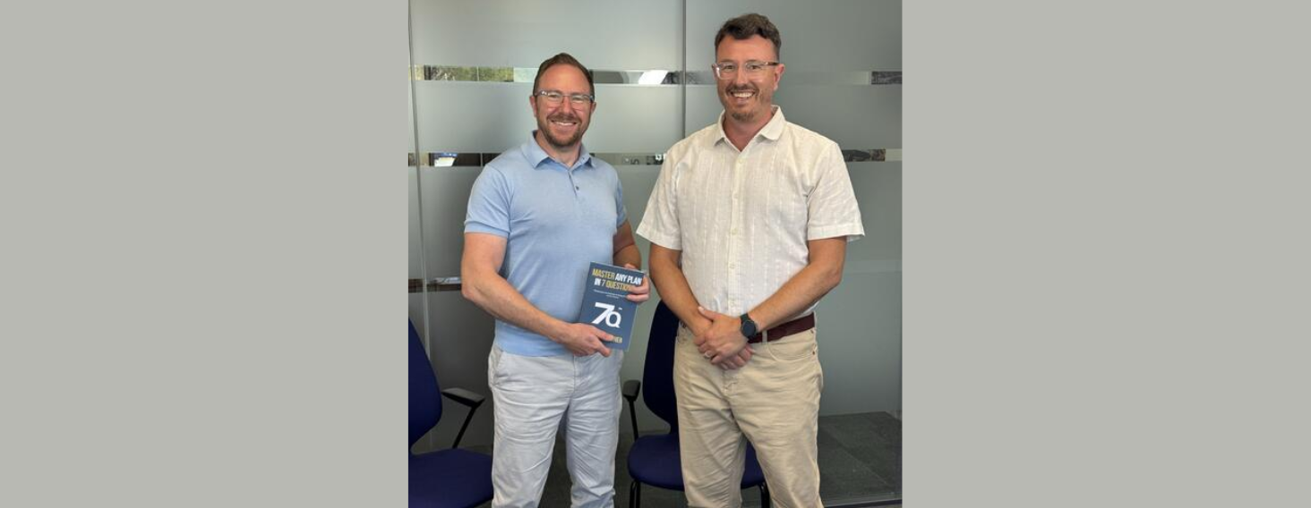 Two men in a casual office pose, one in a light blue polo holding a blue book while the other stands beside him, both smiling in front of frosted glass panels.