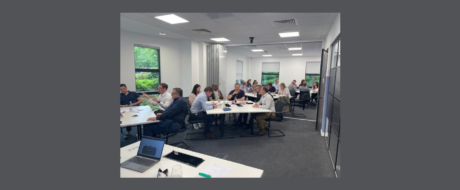 Group of professionals seated around long white tables in a bright office, discussing and taking notes.