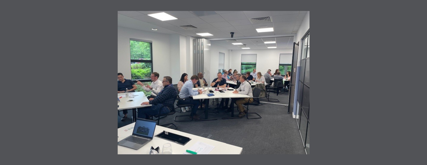 Group of professionals seated around long white tables in a bright office, discussing and taking notes.