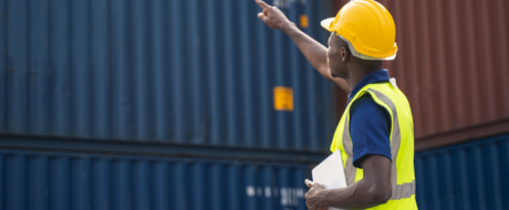 Construction worker in a yellow hard hat and high-visibility vest points toward stacked shipping containers while holding a clipboard in an outdoor yard.