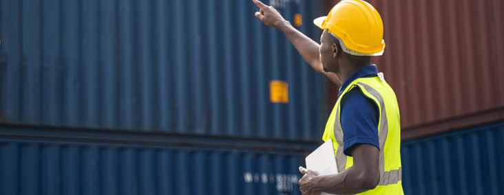 NEW - News item size 82 | Thames Valley Chamber of Commerce Construction worker in a yellow hard hat and high-visibility vest points toward stacked shipping containers while holding a clipboard in an outdoor yard.