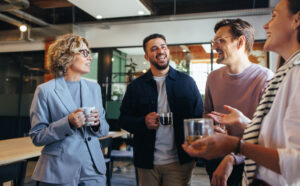 Four coworkers stand at a modern office cafe, smiling and holding cups and glasses, engaged in conversation.