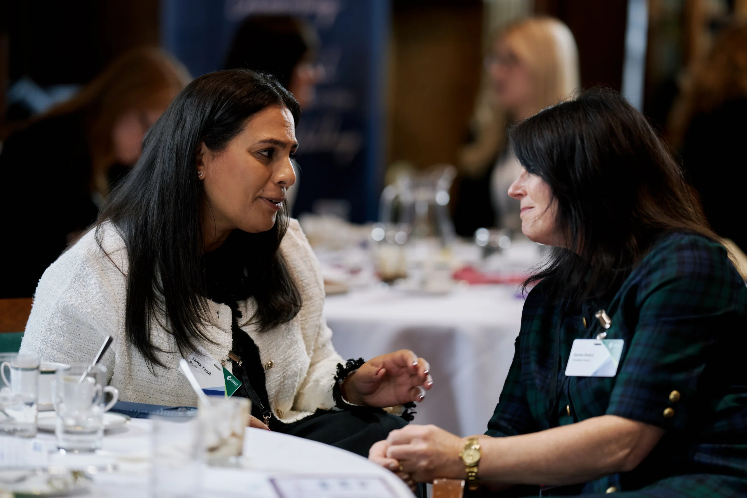 Two women talk across a round dining table at an event, with cups and glassware visible in the foreground.