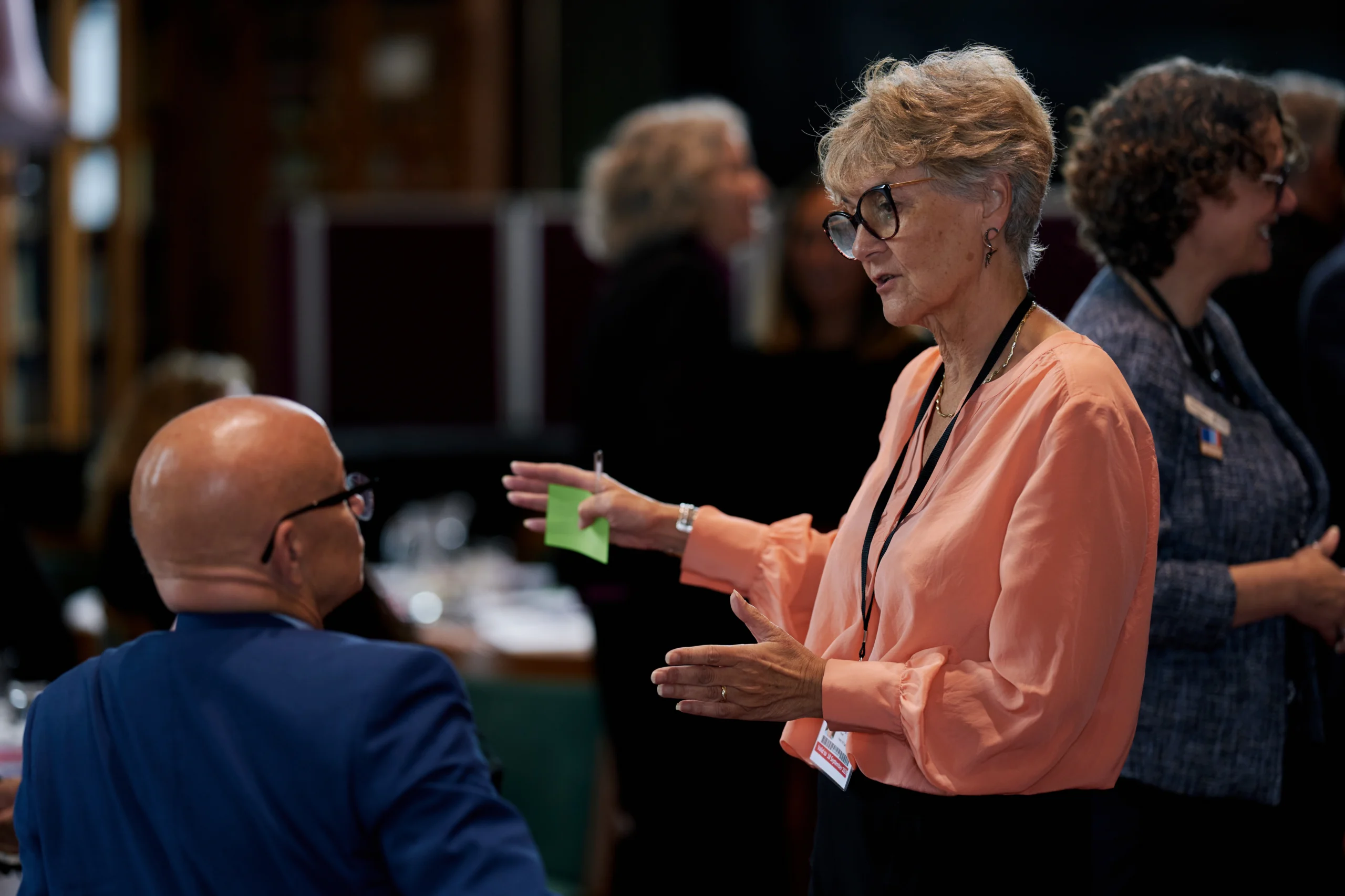 Woman in a peach blouse hands a sticky note to a seated man at a conference or workshop.